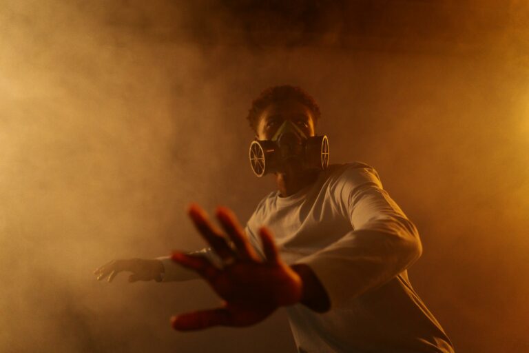 A man in a gas mask reaches out through a smoky, backlit haze, suggesting pollution or danger.